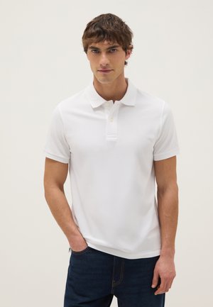 Giovane uomo con capelli corti castani che indossa una polo bianca a maniche corte semplice e jeans blu scuro, in piedi contro uno sfondo chiaro.