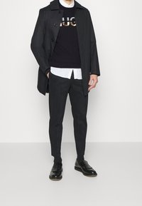 Cappotto nero su un maglione scuro con logo metallico, camicia bianca e pantaloni neri. Texture lisce e mocassini neri lucidi.