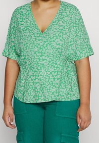 Top floral verde con mangas cortas, que presenta un escote en V y un lazo lateral. El diseño incluye pequeñas margaritas blancas sobre una tela texturizada.