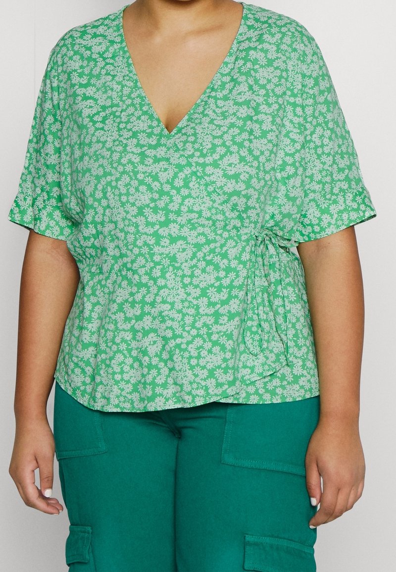 Top floral verde con mangas cortas, que presenta un escote en V y un lazo lateral. El diseño incluye pequeñas margaritas blancas sobre una tela texturizada.