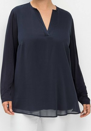 Kvinde iført en løs, langærmet navyblå bluse med V-formet udskæring, kombineret med hvide bukser mod en ensfarvet baggrund.