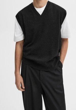 Mężczyzna ubrany w czarny bezrękawnik z dekoltem w serek na białej, krótkorękawowej koszulce, czarne spodnie, stoi z jedną ręką w kieszeni.