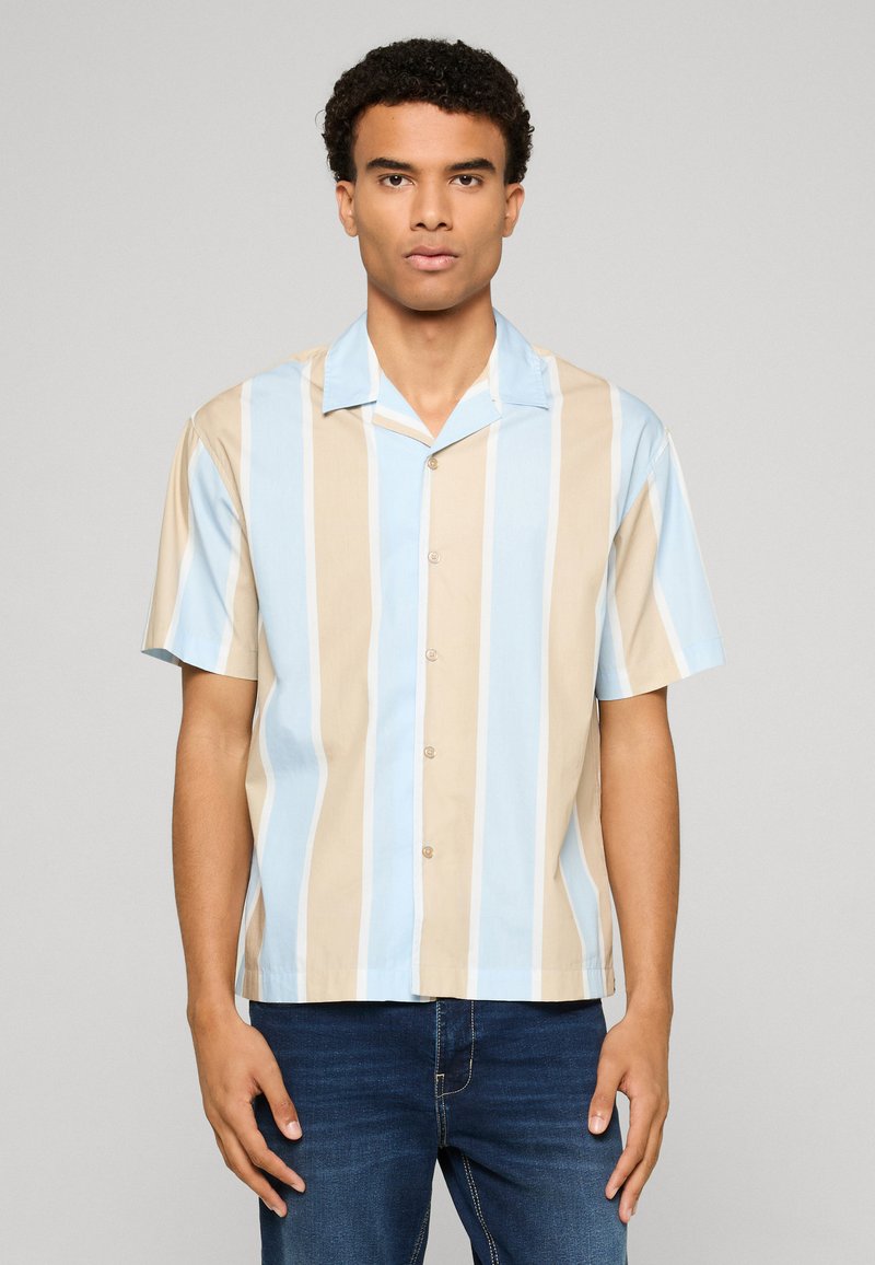 Jeune homme portant une chemise à manches courtes boutonnée avec des rayures verticales bleues, beiges et blanches, associée à un jean bleu foncé.