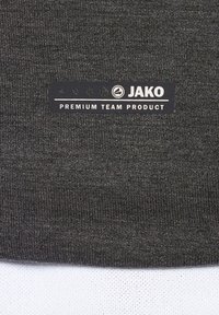 Tissu de maillot de sport gris foncé avec une surface texturée. Présente une étiquette noire avec l'inscription "JAKO PREMIUM TEAM PRODUCT" près du bord inférieur.