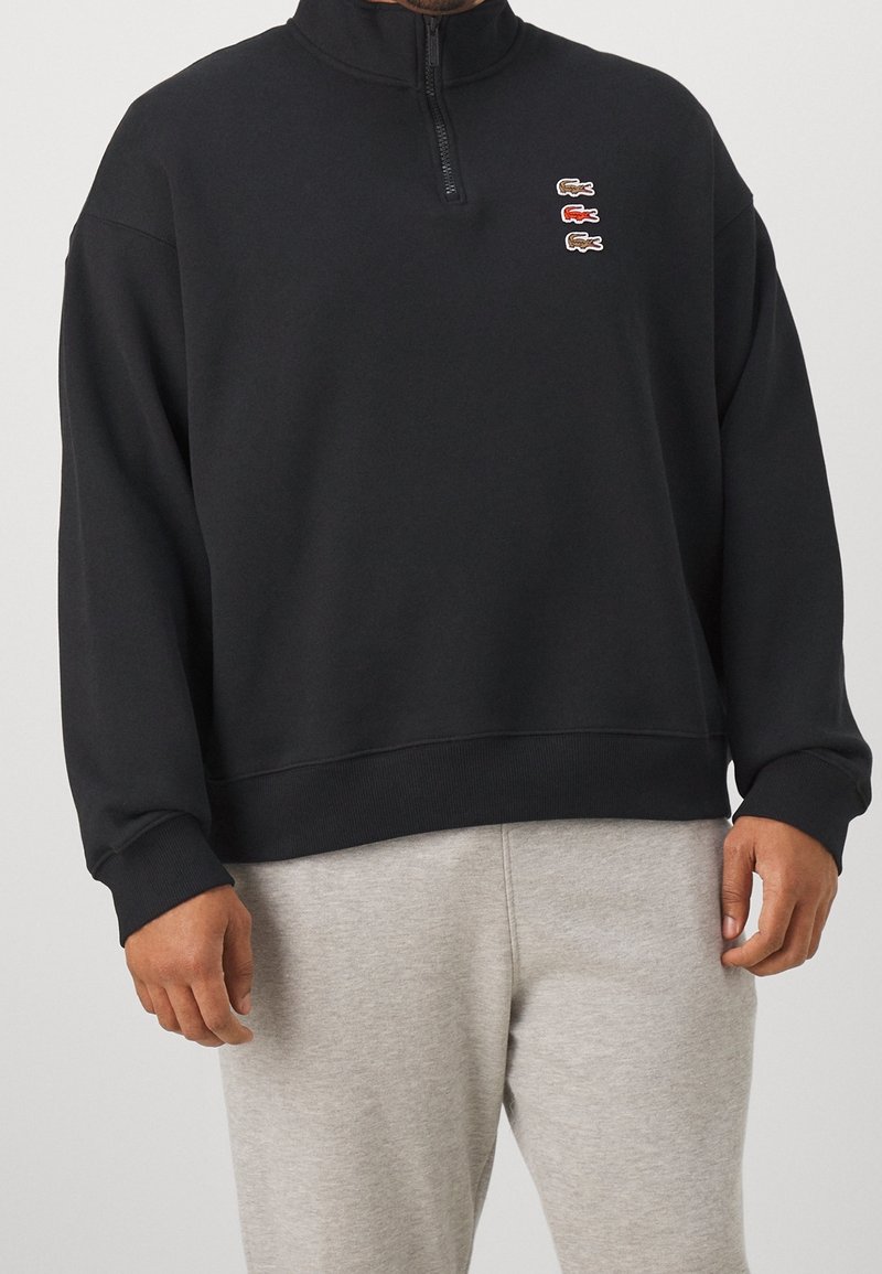 Homme portant un sweat-shirt noir à fermeture éclair sur le quart supérieur, avec trois petits logos crocodile sur la poitrine et un pantalon de jogging gris clair.