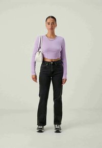 Woman standing wearing a lavender cropped long-sleeve top, black high-waisted jeans, sneakers, gold necklace and earrings, carrying a white shoulder bag.