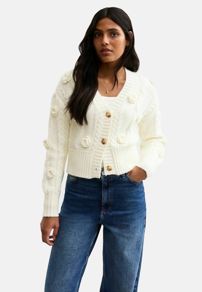 Cardigan bianco a maglia con accenti floreali testurizzati, polsini a coste e tre bottoni in legno, abbinato a jeans blu a vita alta.