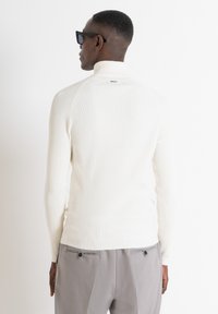 Maglione a collo alto bianco con una vestibilità aderente, caratterizzato da un colletto alto e una texture a costine, abbinato a pantaloni grigi.