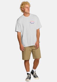 Camiseta blanca de algodón con un gráfico de montaña colorido en el pecho, combinada con pantalones cortos caqui y zapatillas altas negras.