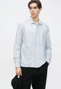 Jaunas vyras dėvi šviesiai mėlyną dryžuotą marškinių su sagutėmis ir juodas kelnes, kairėje rankoje laiko juodą kepurę.