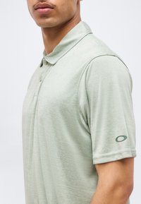 Άνδρας που φοράει ανοιχτοπράσινο polo με κοντά μανίκια, γιακά και δύο κουμπιά, με το λογότυπο Oakley στο αριστερό μανίκι, ουδέτερο φόντο.