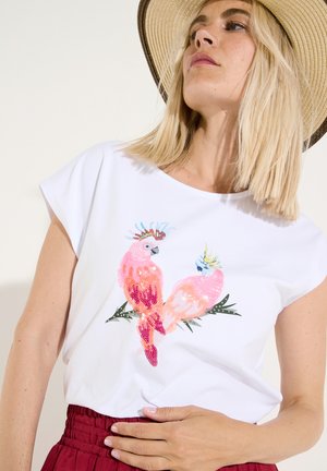Femme portant une chemise blanche avec deux oiseaux cacatoès roses colorés brodés sur le devant, un chapeau beige à large bord et un pantalon rouge.