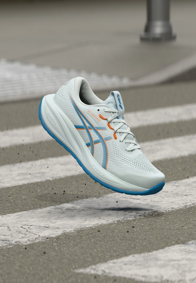 Λευκό και μπλε αθλητικό παπούτσι με πορτοκαλί λεπτομέρειες στον αέρα πάνω από διάβαση πεζών, παρουσιάζοντας λεπτομερή σόλα και ανθεκτικό διχτυωτό επάνω μέρος.