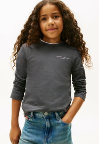 Mädchen mit lockigem Haar, das ein dunkelgraues Tommy Hilfiger Langarmshirt und eine blaue Jeans trägt, mit der Hand in der Tasche, sanft lächelnd vor einem schlichten Hintergrund.
