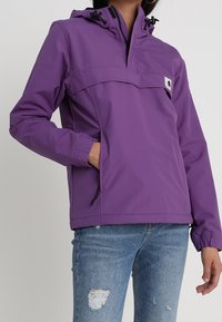 Lilla pullover-jakke med hætte, forlommelomme med lynlås, elastiske manchetter og snorejusteringer. Kombineret med slidte blå jeans.