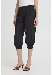 Vrouw draagt zwarte cropped broek met gerimpelde tailleband en boorden, gecombineerd met bruine sandalen met open teen en hak en een witte top.