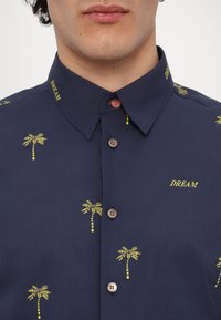 Homme portant une chemise bleu marine avec des imprimés de palmiers jaunes et le mot "DREAM" brodé, bouton rose du col supérieur fermé.