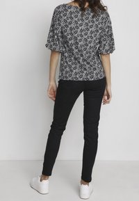 Donna vista da dietro che indossa una maglietta a righe bianche e nere, jeans neri e sneakers bianche, in piedi contro uno sfondo chiaro e uniforme.