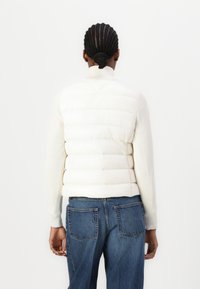 Puffer vest i off-white, med vandret quiltning og ribbede ærmer. Sammen med blå denim jeans, set fra bagenden på en hvid baggrund.