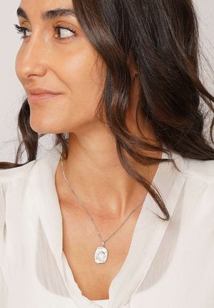 Silberne Anhänger-Halskette an einer feinen Kette, mit einem strukturierten, unregelmäßig geformten Charm, getragen zu einer hellen, fließenden Bluse.