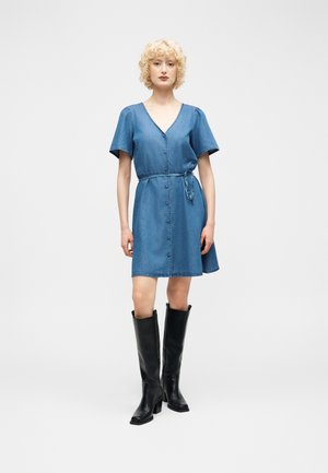 Kvinde stående iført en kortærmet denimkjole med knapper foran og bindebånd i taljen, kombineret med knæhøje sorte støvler mod en hvid baggrund.