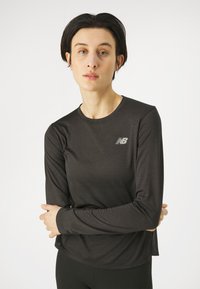 Mustassa pitkähihaisessa urheilupaidassa on tekstuuroitu kangas, pyöreä kaula-aukko ja pieni heijastava New Balance -logo rinnassa.