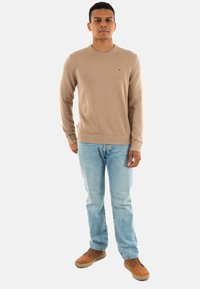 Maglione beige lavorato a maglia con orlo a costine, abbinato a jeans azzurri chiari e scarpe marroni, in piedi davanti a uno sfondo bianco.