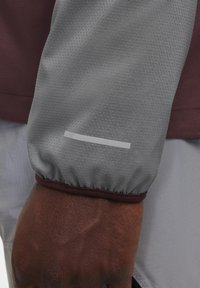 Dunkelhäutige Hand, die einen grauen Jackenärmel mit gepunkteter Textur und einem reflektierenden Streifen oberhalb des Handgelenks trägt.