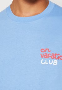 Ljusblå bomull T-shirt med rund halsringning. Texten "on vacation CLUB" är tryckt på bröstet i röd och vit.