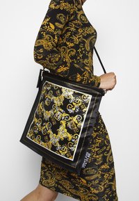 Svart tote-väska med en blank finish, prydd med guld- och vita barockmönster, i kvadratisk design, samt en logodetalj längs kanten.