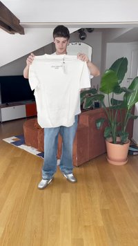 Ung mann holder en hvit T-skjorte med prislapp i en moderne stue med tregulv, brun sofa, stor grønn plante og TV i bakgrunnen.