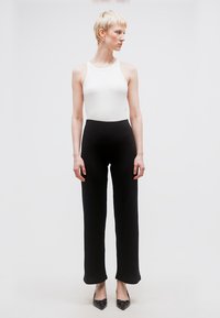 Άτομο με κοντά ξανθά μαλλιά, φορώντας λευκό αμάνικο τοπ, μαύρο παντελόνι ψηλόμεσο και μαύρα μυτερά παπούτσια, στέκεται σε απλό φόντο.