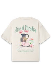 Kremfarget t-skjorte med løs passform, prydet med et fargerikt motiv av en mugge, frukt og blomster. Teksten sier "Slice of Paradise" i mintgrønn.