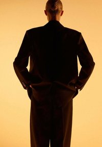 Traje negro de chaqueta y pantalones, tela suave, pose de pie, contra un fondo naranja cálido, mostrando la vista trasera con las manos relajadas.