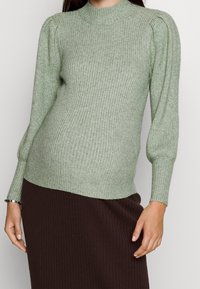 Zielony sweter z prążkowanego dzianiny, z wysokim kołnierzem, bufiastymi ramionami i długimi rękawami, noszony z brązową spódnicą z prążkowanej dzianiny.