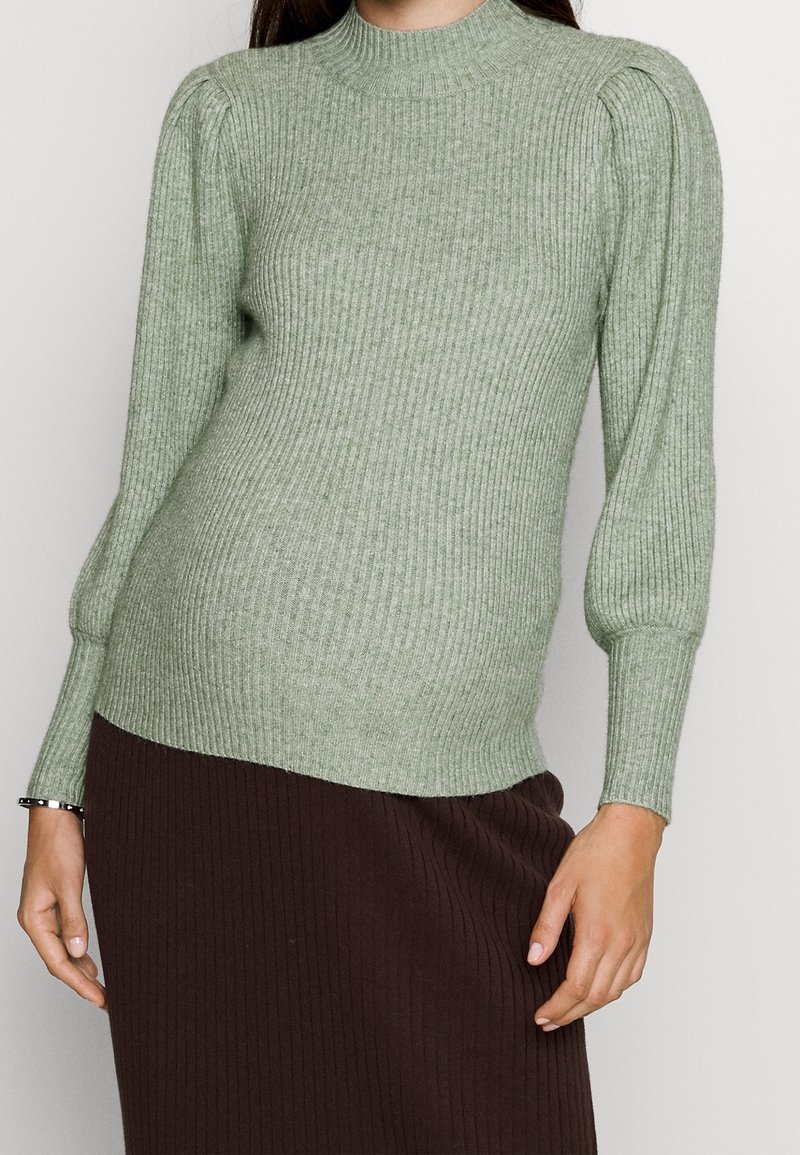 Zielony sweter z prążkowanego dzianiny, z wysokim kołnierzem, bufiastymi ramionami i długimi rękawami, noszony z brązową spódnicą z prążkowanej dzianiny.