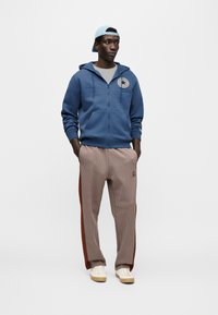 Μπλε φούτερ με φερμουάρ και κυκλικό έμβλημα, γκρι μπλούζα, καρό καφέ παντελόνια με κόκκινες πλαϊνές ρίγες και ανοιχτόχρωμα αθλητικά παπούτσια.