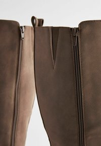 Bottes en cuir marron avec fermetures éclair latérales visibles et languettes cousues, présentées en gros plan sur un fond neutre.