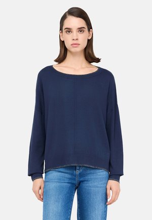 Donna con capelli castano lisci, che indossa un maglione a maniche lunghe blu scuro e jeans azzurri, in piedi davanti a uno sfondo bianco semplice.