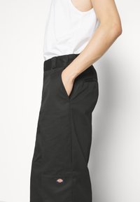 Black pants with a smooth texture, featuring a fitted waistband, side pockets, and a small logo patch on the front. Simple design.