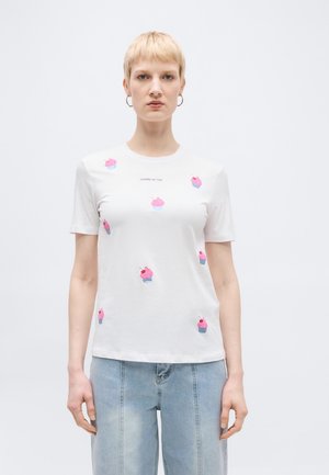 Osoba ubrana w białą koszulkę z różowymi wzorami babeczek i napisem "CHERRY ON TOP", zestawioną z jasnoniebieskimi dżinsami na jednolitym tle.