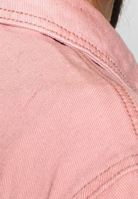 Ljusrosa skjorta i tyg med en strukturerad yta och synlig sömnad längs kragen; delvis synlig axelområde.