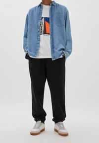 Denim skjorta med knappar i ljusblått över en vit t-shirt med grafiskt tryck, parat med svarta sweatpants och vita sneakers med bruna sulor.