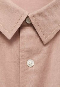 Svetlo roza lanena srajca z ovratnikom na gumb, ki ima bele gumbe in teksturirano površino. Zaslon blizu podrobnosti šivanja.