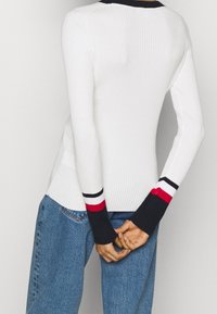 Person, der einen figurbetonten weißen Rippsweater mit schwarzen, weißen und roten gestreiften Manschetten trägt, kombiniert mit blauen Jeans, die Hände hinter dem Rücken verschränkt.