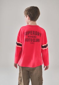 Rød langærmet skjorte med "Superdry Riders Moto Club" print på ryggen. Har sorte og gule striber på ærmerne. Bomulds materiale.
