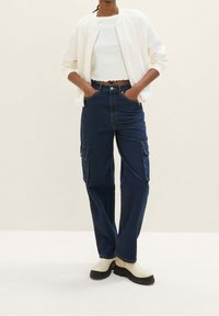 Pantaloni cargo blu scuro con tasche laterali, vita alta e cuciture a contrasto, abbinati a un top corto bianco e stivali color crema.