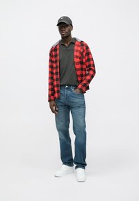 Camisa xadrez vermelha e preta sobre um polo cinza, jeans azuis e ténis brancos; boné cinza com logótipo; roupa casual com camadas de texturas.