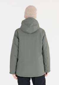 Armeegrüne, wasserdichte Jacke mit Kapuze, die eine glatte Textur und eine lockere Passform aufweist. Eine gestrickte beigefarbene Mütze vervollständigt den Look.
