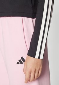 Črno dolgi rokavi s belimi črtami na rokavih, v paru z roza hlačami, ki imajo črni logotip na stegnu, gladka bombažna tkanina.